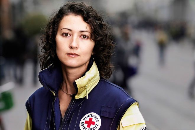 Foto: Eine junge Frau mit dunklen Locken blickt in die Kamera. Sie trägt eine Rotkreuzjacke und wirkt insgesamt sehr verantwortungsbewusst.